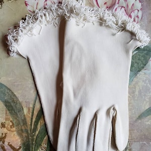 May include: A pair of white cotton gloves with a white lace trim. The gloves are on a floral background.