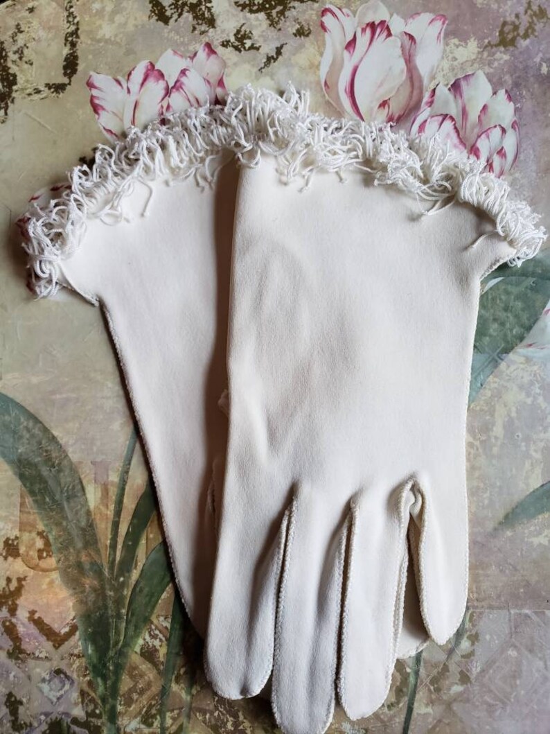 May include: A pair of white cotton gloves with a white lace trim. The gloves are on a floral background.