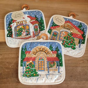 May include: Set of three Christmas-themed pot holders. Each square pot holder features a different shop: a toy store, a candy store, and a bakery, all decorated for the holidays. They have a blue background with falling snow and a wooden ring for hanging.