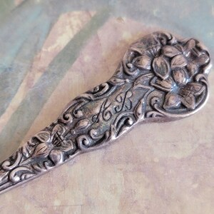 May include: Close-up of a silver spoon with ornate floral and leaf detailing. The spoon's handle features a flower design and the initials "A&B". The spoon has a vintage, antique appearance.