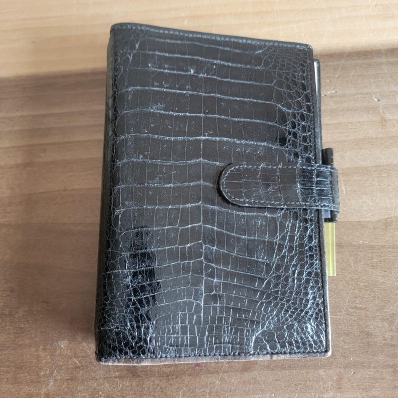 Puede incluir: Una agenda de cuero negro con un patr&oacute;n de cocodrilo en relieve y un portaplumas dorado.