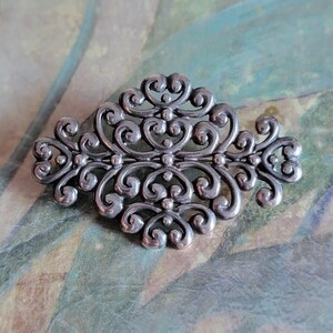 May include: Silver-toned filigree brooch with an ornate, symmetrical design. The brooch features swirling, scroll-like patterns and heart-shaped details. The metal has a slightly aged appearance, adding to its vintage charm. The brooch is a decorative accessory.