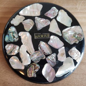 May include: A round, black coaster inlaid with iridescent abalone shells and white shell fragments. A small metal tag reads "HAWAII". The coaster is set on a wooden surface.