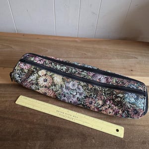Puede incluir: Un estuche de tapiz floral con dos cremalleras negras. La funda es en tonos de rosa, verde y morado. Una regla amarilla con el texto "NEW MATH RULER" est&aacute; al lado de la funda.