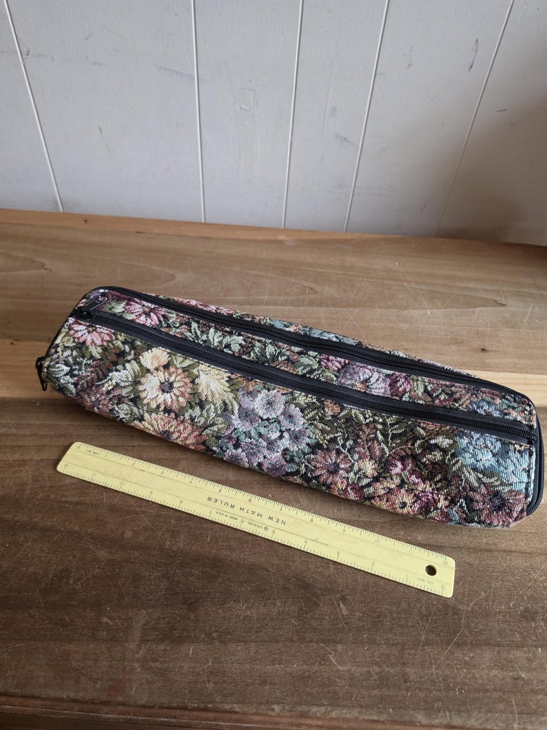 Puede incluir: Un estuche de tapiz floral con dos cremalleras negras. La funda es en tonos de rosa, verde y morado. Una regla amarilla con el texto "NEW MATH RULER" est&aacute; al lado de la funda.