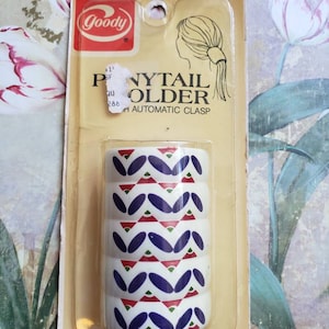 May include: A vintage Goody ponytail holder in its original packaging. The white holder features a repeating pattern of blue, red, and green shapes. The packaging reads "PONYTAIL HOLDER" and "AUTOMATIC CLASP". Made in the USA.