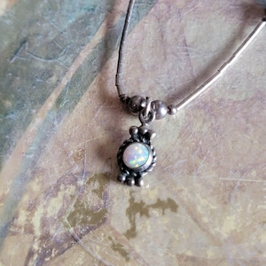 May include: A silver necklace with a small pendant featuring a round, iridescent opal stone. The pendant is framed by a decorative silver border. The necklace has a delicate, beaded chain.