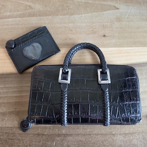 May include: A black rectangular wallet with a crocodile-embossed texture, featuring a braided handle, silver-toned metal hardware, and a zipper closure. Beside it sits a matching black card holder with a heart-shaped cutout window.