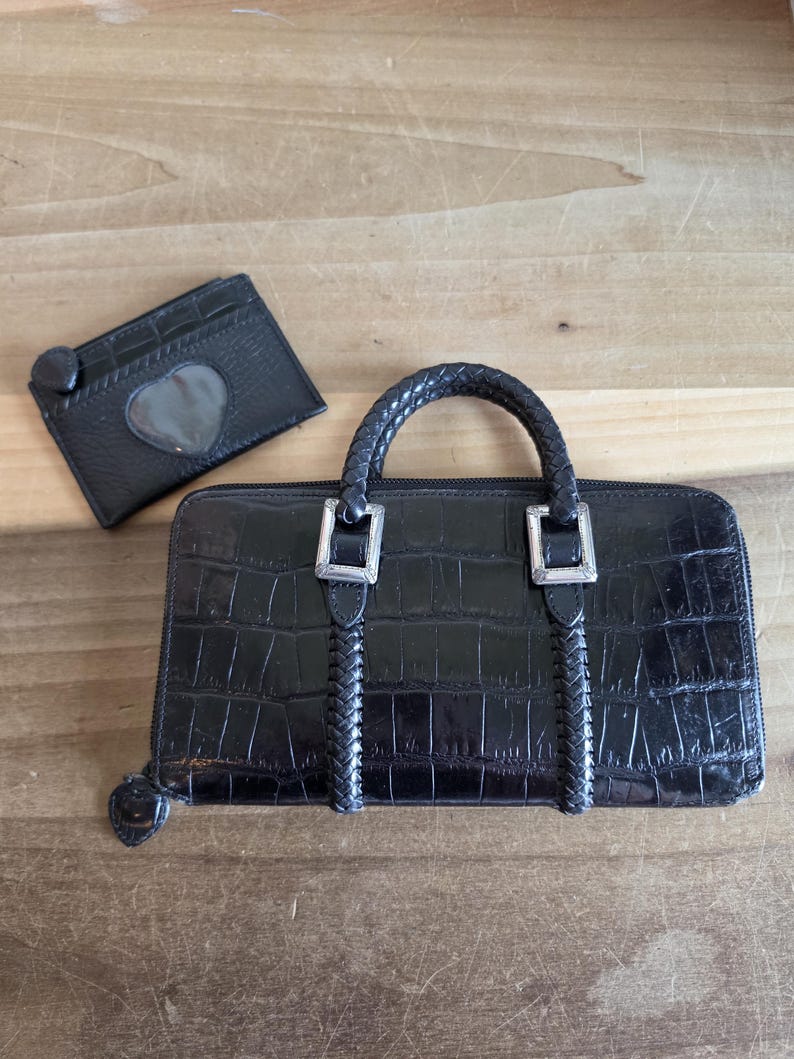 May include: A black rectangular wallet with a crocodile-embossed texture, featuring a braided handle, silver-toned metal hardware, and a zipper closure. Beside it sits a matching black card holder with a heart-shaped cutout window.