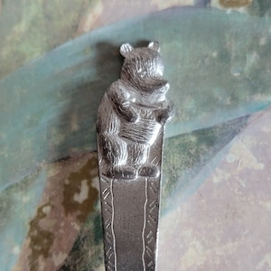 May include: Silver baby spoon with a bear design. The bear is sitting on its haunches with its paws in front of it.