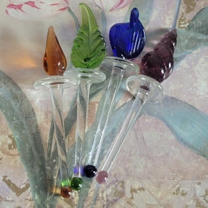 May include: Five glass decorative picks with different colored glass tops. The tops are a green leaf, a blue swirl, a purple shell, a brown flame, and a clear round ball. The picks are on a floral patterned surface.