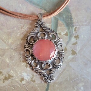 May include: A silver pendant with an ornate design features a large, round, pink gemstone. The pendant is attached to a brown leather cord.
