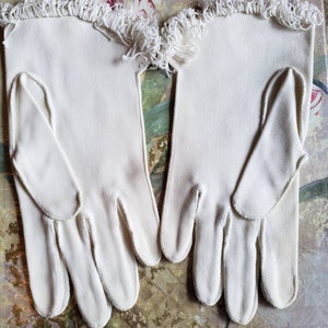 May include: A pair of white cotton gloves with a white fringe trim. The gloves are laying flat on a floral patterned surface.