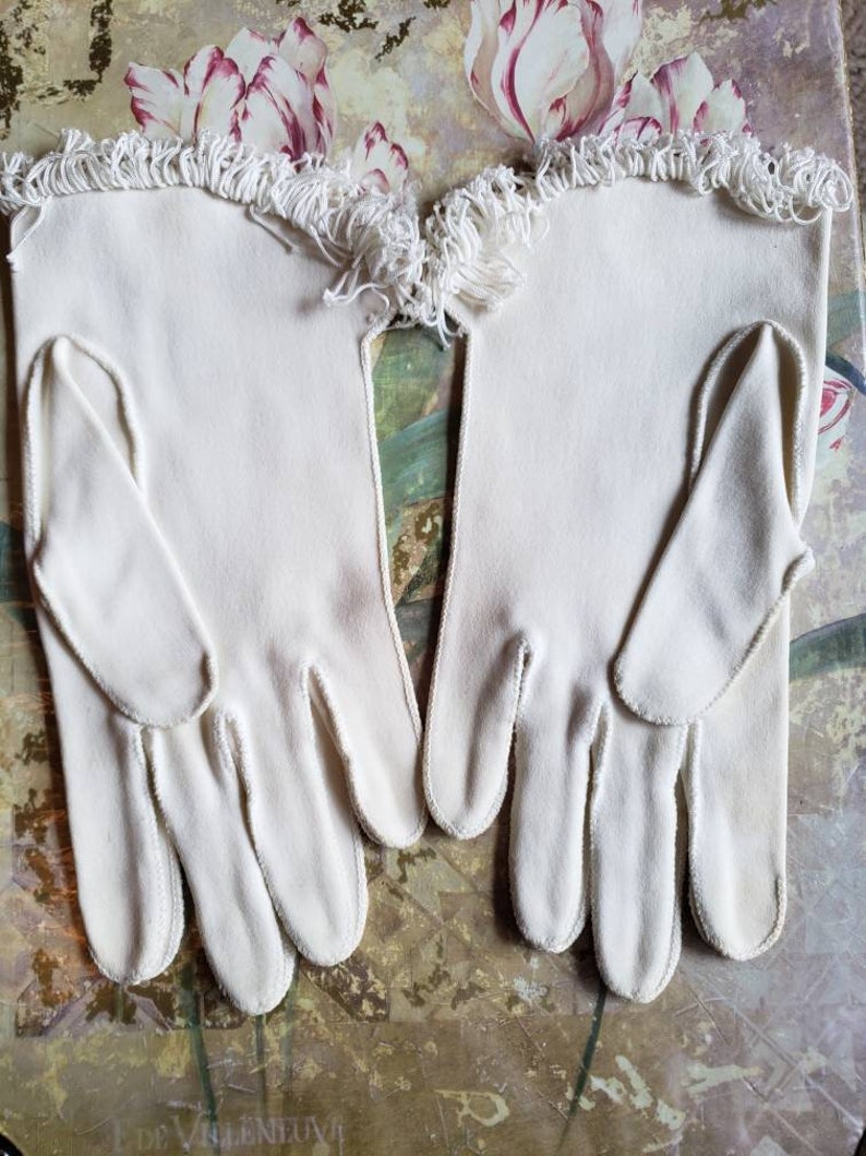 May include: A pair of white cotton gloves with a white fringe trim. The gloves are laying flat on a floral patterned surface.