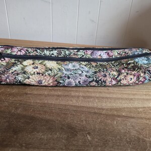 Puede incluir: Un estuche largo y rectangular de tapiz floral con cierre de cremallera negro. El estuche presenta un estampado de flores coloridas en tonos rosa, amarillo y azul sobre un fondo oscuro. El estuche est&aacute; sobre una superficie de madera.