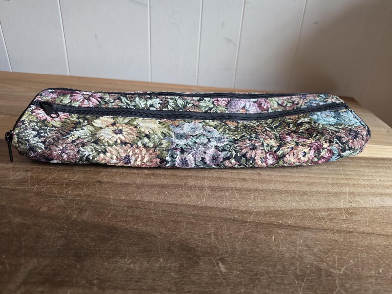 Puede incluir: Un estuche largo y rectangular de tapiz floral con cierre de cremallera negro. El estuche presenta un estampado de flores coloridas en tonos rosa, amarillo y azul sobre un fondo oscuro. El estuche est&aacute; sobre una superficie de madera.