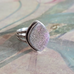 May include: A silver ring with a sparkly, iridescent, light purple and white gemstone. The ring has a triple band design.