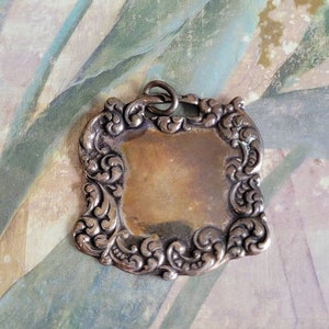 May include: A silver pendant with an ornate, scrolled frame and a blank, smooth surface in the center. The pendant has a loop at the top for attaching a chain.