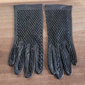 May include: A pair of black leather gloves with a woven pattern on the back of the hand. The gloves have a classic design with individual fingers and a smooth texture. The gloves are displayed on a wooden surface.
