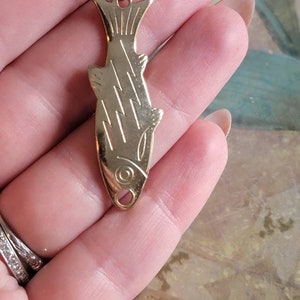 May include: A gold-toned fish pendant necklace. The pendant is detailed with etched lines and a small loop at the top for attaching a chain.