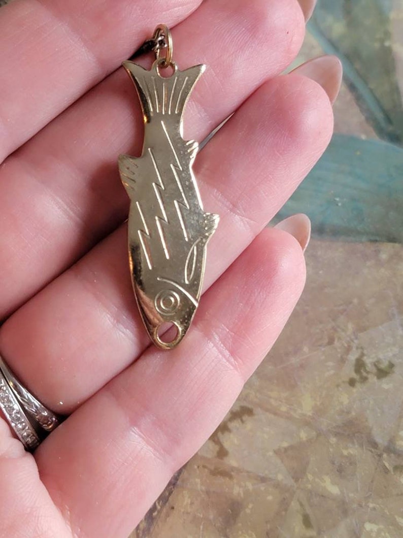 May include: A gold-toned fish pendant necklace. The pendant is detailed with etched lines and a small loop at the top for attaching a chain.