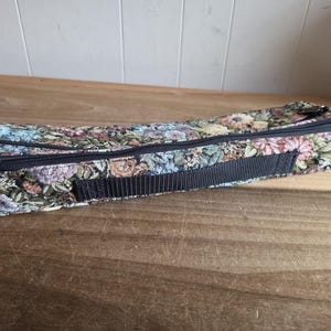 Puede incluir: Un estuche rectangular de tapiz floral con cremallera negra y correa tejida negra. El estuche presenta un colorido estampado de flores en tonos azules, rosas, verdes y marrones. El estuche est&aacute; cerrado y descansa sobre una superficie de madera.