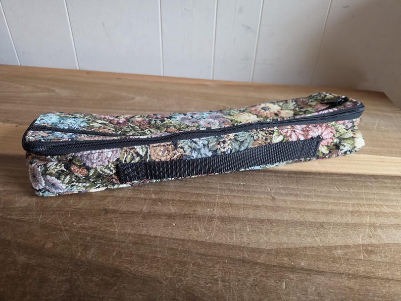 Puede incluir: Un estuche rectangular de tapiz floral con cremallera negra y correa tejida negra. El estuche presenta un colorido estampado de flores en tonos azules, rosas, verdes y marrones. El estuche est&aacute; cerrado y descansa sobre una superficie de madera.