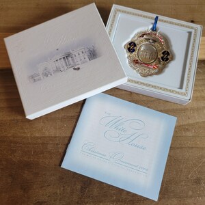 May include: A White House Christmas ornament from 2006, displayed in its original box. The ornament is gold-toned with red, white, and blue accents. The box and accompanying booklet feature the White House and the text "Christmas Ornament 2006."