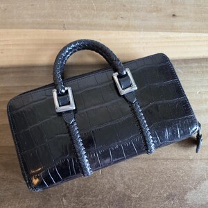 May include: A black rectangular wallet with a crocodile-embossed texture, featuring a braided top handle and silver-toned metal hardware accents. The wallet includes a zipper closure and braided detailing that extends vertically from the handle base.