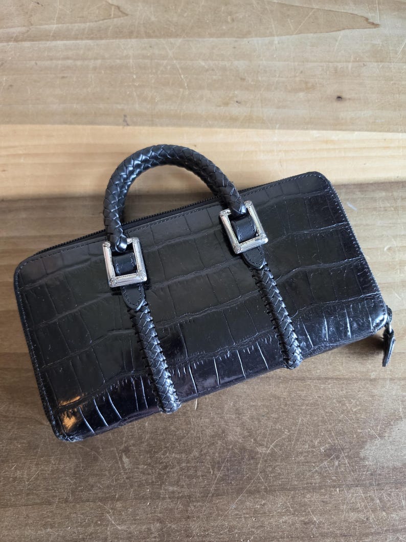 May include: A black rectangular wallet with a crocodile-embossed texture, featuring a braided top handle and silver-toned metal hardware accents. The wallet includes a zipper closure and braided detailing that extends vertically from the handle base.