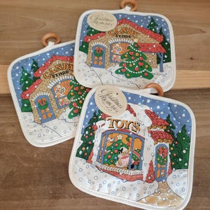 May include: Three square Christmas pot holders with a winter scene featuring a candy store and a toy store. Each pot holder has a wooden ring for hanging and a tag that reads "Christmas Memories". The pot holders are white with a blue border.