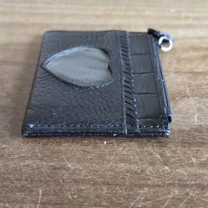 May include: A black leather card holder featuring a heart-shaped clear plastic window on the front, a textured grain finish, a section with a crocodile-embossed pattern, and a silver-toned metal zipper pull attached to the side of the slim wallet.