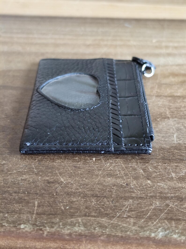 May include: A black leather card holder featuring a heart-shaped clear plastic window on the front, a textured grain finish, a section with a crocodile-embossed pattern, and a silver-toned metal zipper pull attached to the side of the slim wallet.