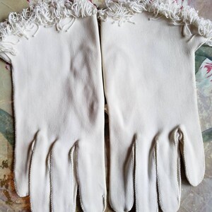 May include: A pair of white cotton gloves with a fringe trim at the wrist.