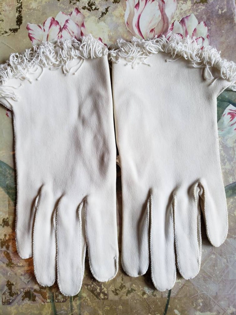 May include: A pair of white cotton gloves with a fringe trim at the wrist.
