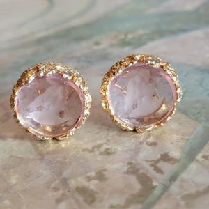 May include: A pair of gold-toned stud earrings with a textured, organic design. Each earring features a light pink, translucent stone with an etched design. The earrings are set against a soft, neutral background.