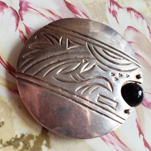 May include: A silver circular brooch with an intricate design of stylized leaves and a black onyx stone.