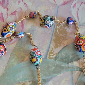 May include: A gold chain necklace with colorful, floral patterned glass beads. The beads are oval shaped and have a variety of colors, including red, blue, green, yellow, and white.