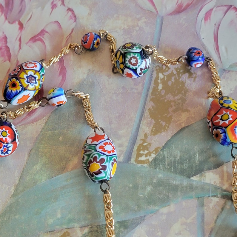 May include: A gold chain necklace with colorful, floral patterned glass beads. The beads are oval shaped and have a variety of colors, including red, blue, green, yellow, and white.