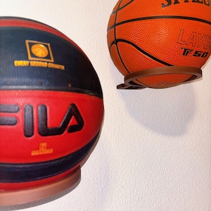 Può includere: Due palloni da basket, uno arancione e uno rosso, montati su un muro bianco. La palla arancione ha la scritta "SPALDING". La palla rossa ha la scritta "FILA". Entrambe le palle sono in supporti marroni.
