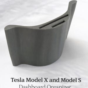 Pode incluir: Um organizador de painel cinza para veículos Tesla Model X e Model S. O organizador tem um design curvo com duas ranhuras retangulares. O texto na imagem diz "Tesla Model X and Model S Dashboard Organizer".