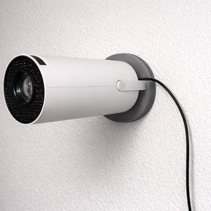 Könnte beinhalten: Eine weiße Überwachungskamera an einer strukturierten weißen Wand befestigt. Die Kamera hat einen zylindrischen Körper mit einer schwarzen Netzfront und einer grauen Halterung. Ein schwarzes Kabel erstreckt sich von der Kamera.