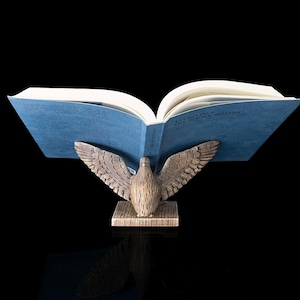 Pode incluir: Um livro aberto com capas azuis é suportado por uma escultura de águia de madeira. A águia tem as asas abertas e está pousada sobre uma base retangular. As páginas do livro são brancas e o fundo é preto.