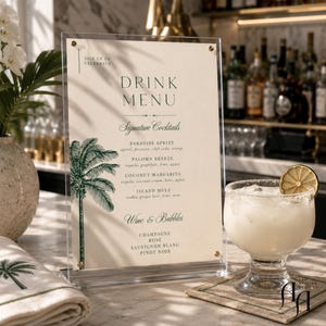 May include: A vertical acrylic sign holder displaying a cream-colored drink menu with dark green text and a palm tree illustration. The menu lists signature cocktails and wine options. A glass of cocktail with a lime slice sits beside it.