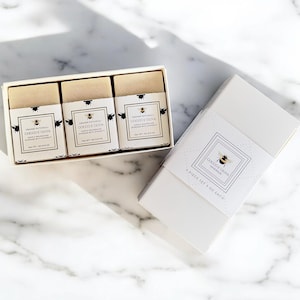 May include: A white box set containing three bars of soap, each in a separate wrapper, and a separate box. The soap wrappers and box feature a bee logo and the text "Orange Patchouli Cocoo e Oliva". The set is on a marble surface.