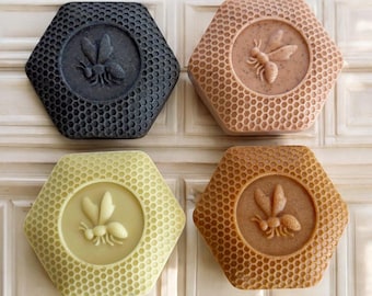 May include: Four hexagon-shaped soaps in shades of black, peach, yellow, and brown. Each soap features a detailed bee design in the center, surrounded by a honeycomb pattern. The soaps are arranged on a white surface.
