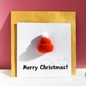 May include: A greeting card with a small, knitted red Santa hat and the text "Merry Christmas!" on a white background. The card is layered over a gold envelope, set against a pink background.
