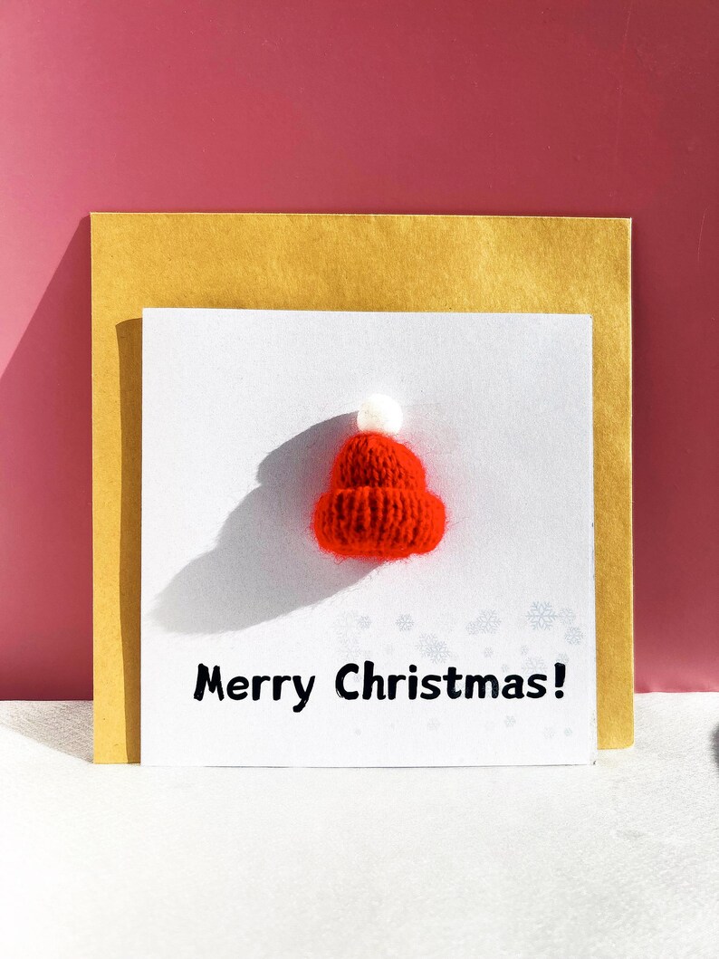 May include: A greeting card with a small, knitted red Santa hat and the text "Merry Christmas!" on a white background. The card is layered over a gold envelope, set against a pink background.
