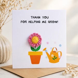 May include: A white greeting card with the text THANK YOU FOR HELPING ME GROW! in black. The card features a 3D pink and yellow crocheted flower in an orange pot and a printed yellow watering can with small blue and pink flower illustrations.