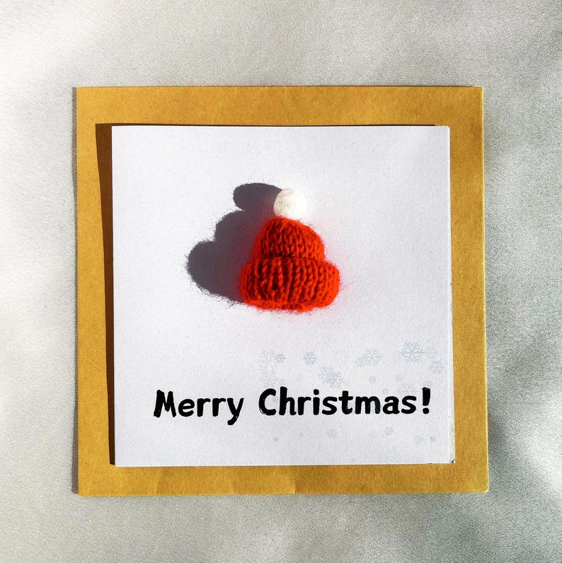 May include: A handmade Christmas card featuring a small, knitted red hat with a white pom-pom. The card is set on a white background, framed by a gold border, and the text "Merry Christmas!" is printed below the hat.